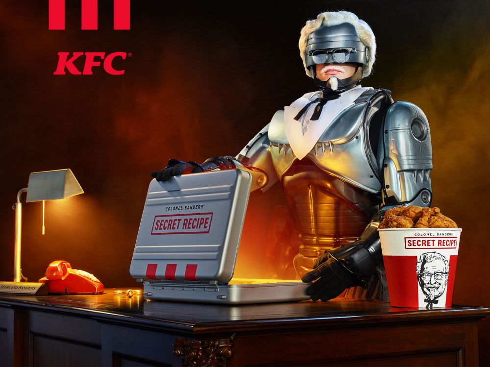 A person in robotic Colonel Sanders armor stands at a desk with a 'Secret Recipe' briefcase and fried chicken, promoting KFC.