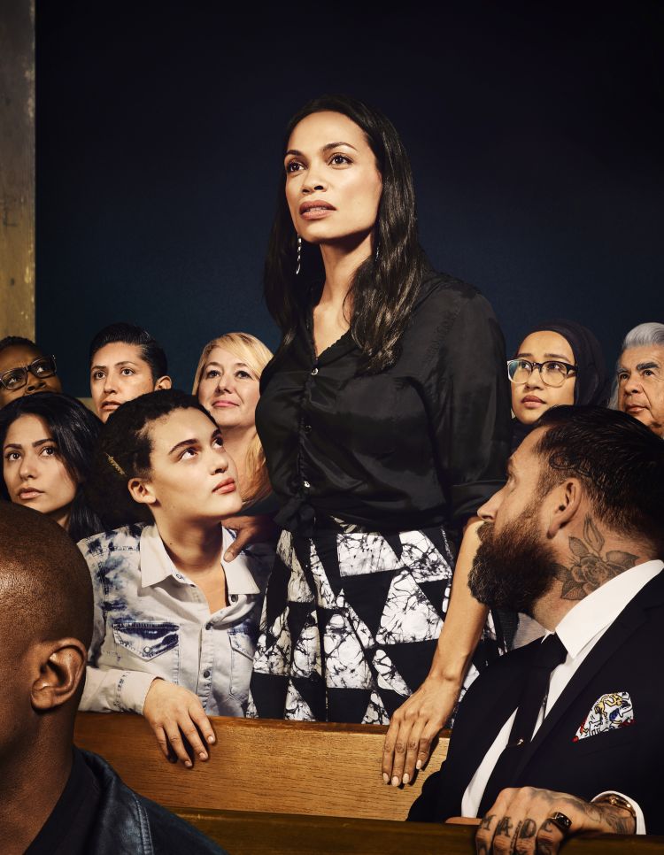Rosario Dawson stands amidst a diverse group of people in an indoor setting, wearing a black blouse and patterned skirt.