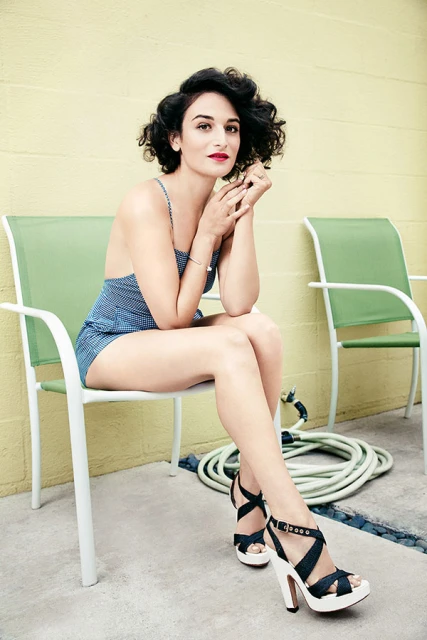 A curly-haired woman poses while sitting on a green chair in front of a beige wall wearing a blue bathing suit and dark platform heels.