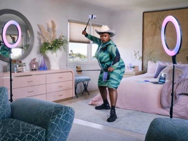 Adult female in a turquoise dress and cowboy hat poses in a bedroom studio with ring lights, cell phones, and a laptop.