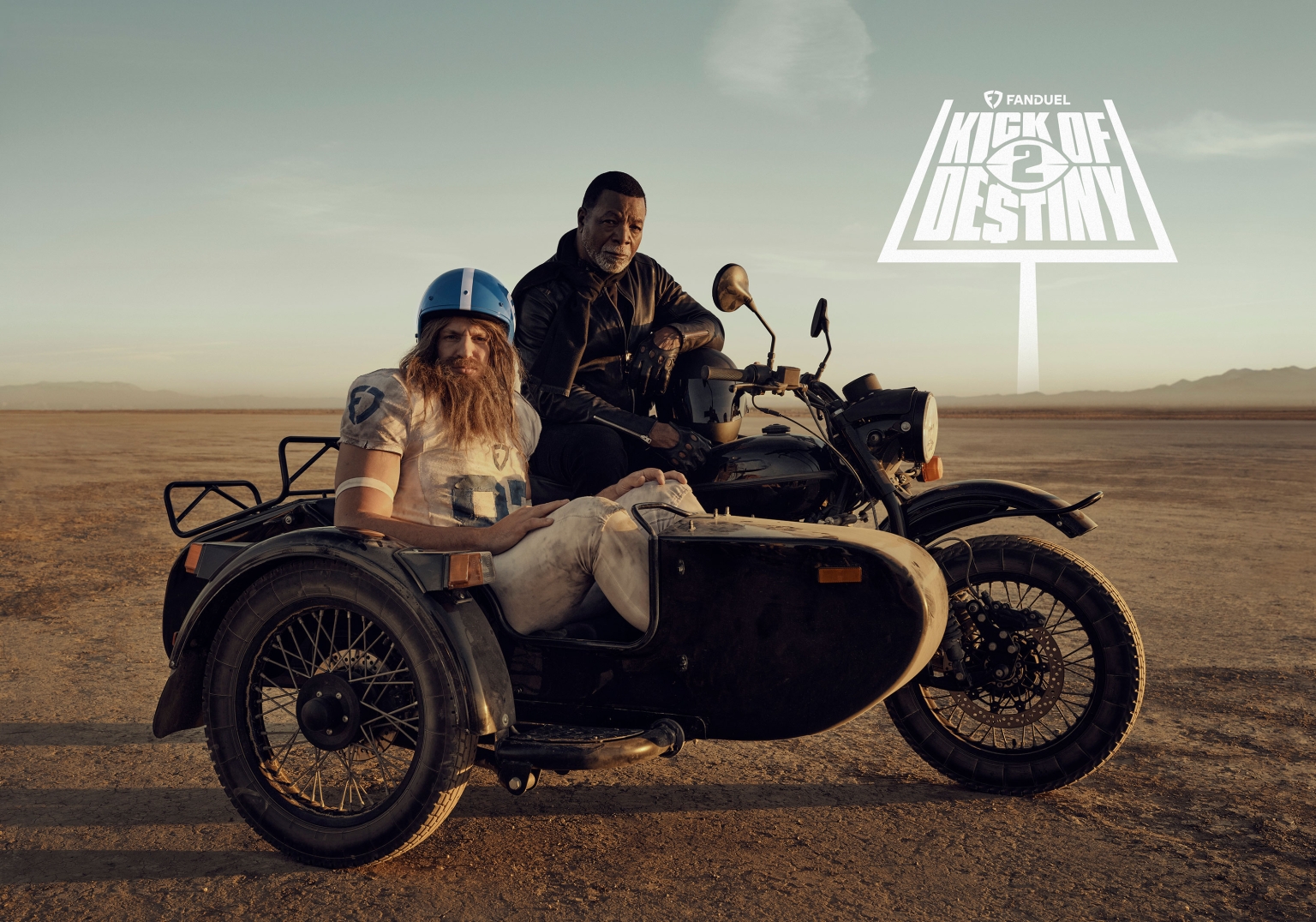 Two men, one in football gear in a sidecar and the other on a motorcycle, are silhouetted against a desert backdrop with a promotional sign.