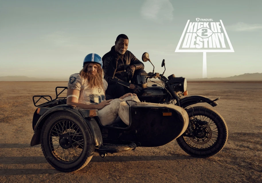 Two men, one in football gear in a sidecar and the other on a motorcycle, are silhouetted against a desert backdrop with a promotional sign.