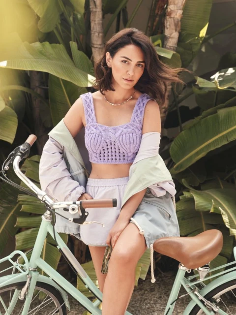A brunette woman sits on a turquoise bicycle in a light purple knit top and shorts, with palm trees in the sunny background.