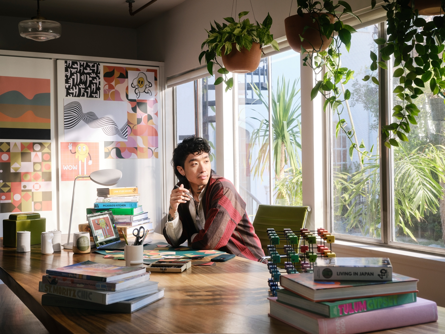 Adult with pen sits at a table in a bright indoor space with books, art, and potted plants near the window, looking to the right.