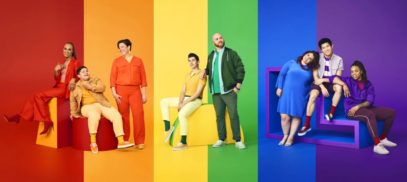 Diverse group of nine people wearing brightly colored clothing posing in front of a split color background of red, orange, yellow, green, blue, and purple.