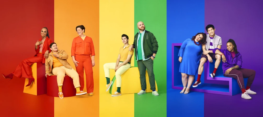 Diverse group of nine people wearing brightly colored clothing posing in front of a split color background of red, orange, yellow, green, blue, and purple.