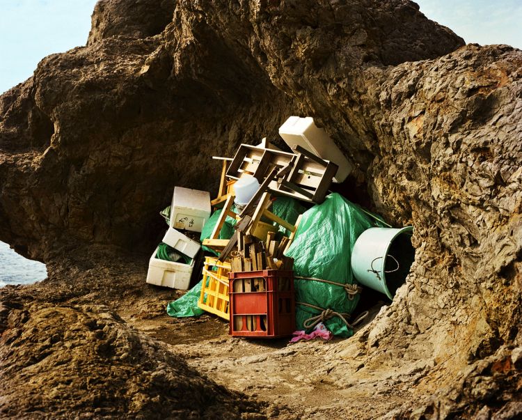 A pile of discarded items, including crates, boxes, and a tarp, rests at the base of a rocky outcropping by the sea under bright daylight.