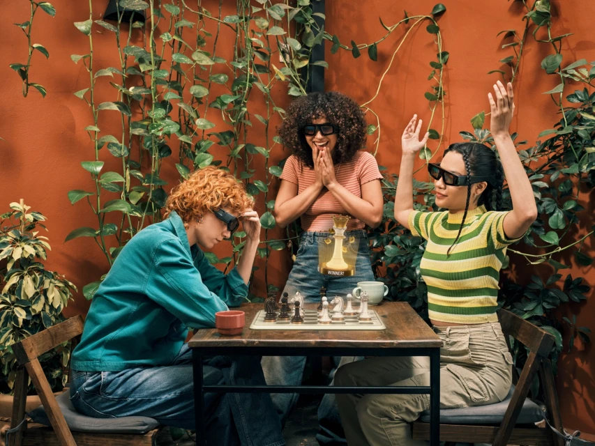 Three young adults in smart glasses playing chess on outdoor patio surrounded by greenery and terracotta walls, enjoying a casual game together.