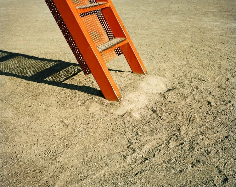 Orange playground slide stairs in a sandy park with visible tire and shoe tracks, creating a playful and textured outdoor scene.