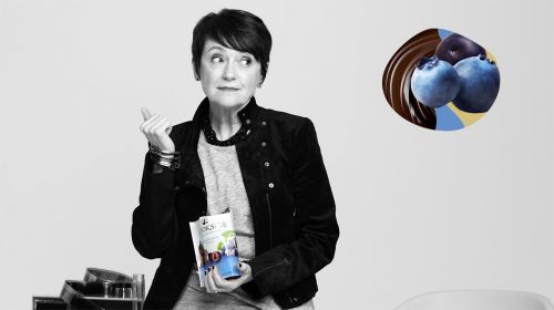 Woman poses indoors with snack pack in front of an advertisement featuring blueberries covered in chocolate, on a monochrome background.