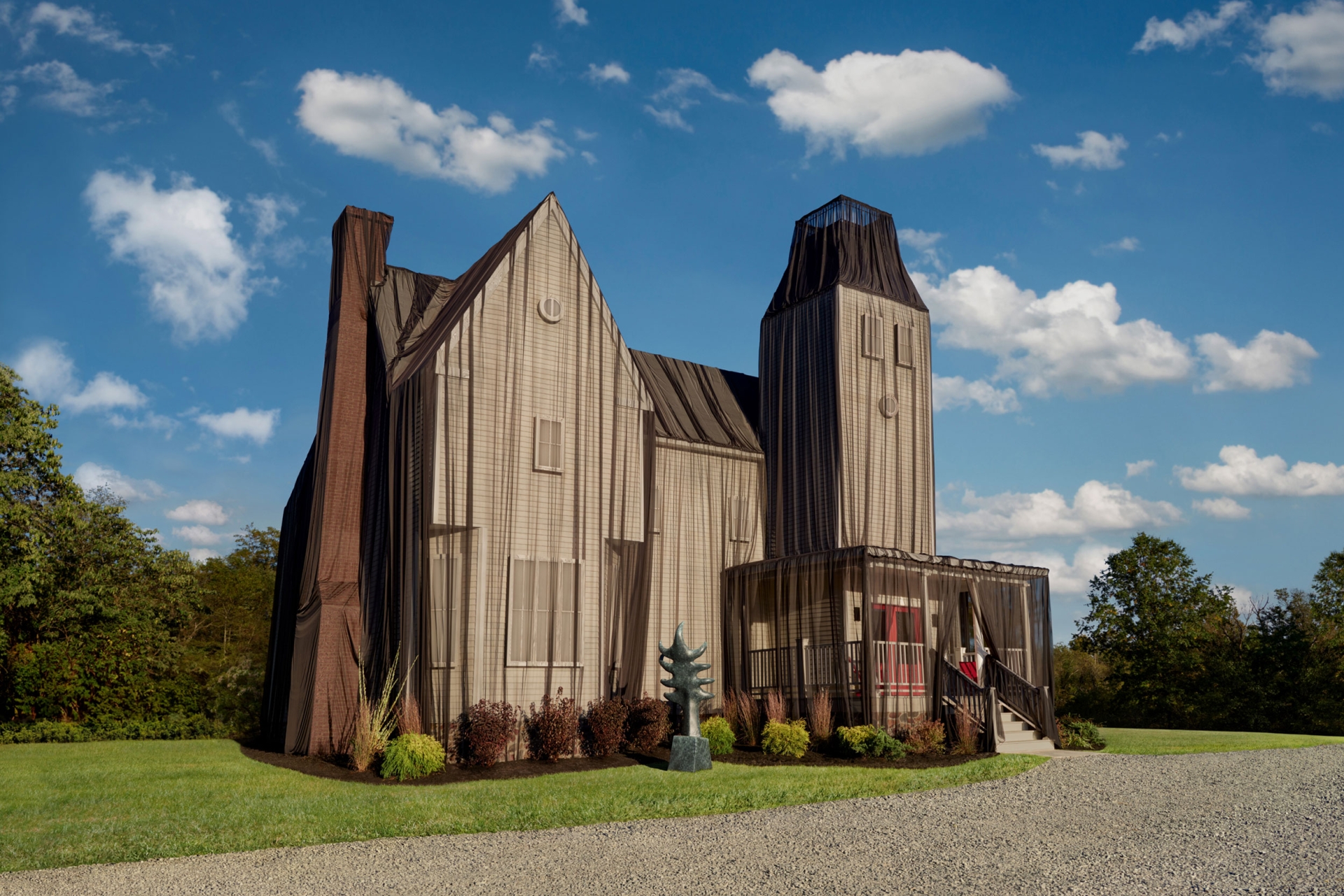 Exterior of a large, light-colored house completely draped in sheer black fabric, standing on a lawn beneath a partly cloudy sky.