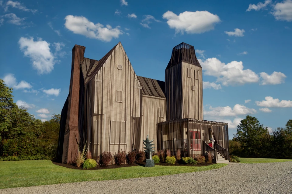 Exterior of a large, light-colored house completely draped in sheer black fabric, standing on a lawn beneath a partly cloudy sky.