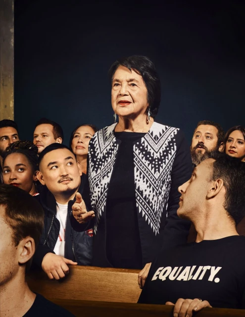 Indoor shot of an older woman in black and white patterned sweater, surrounded by a diverse group of people in a dimly lit space.