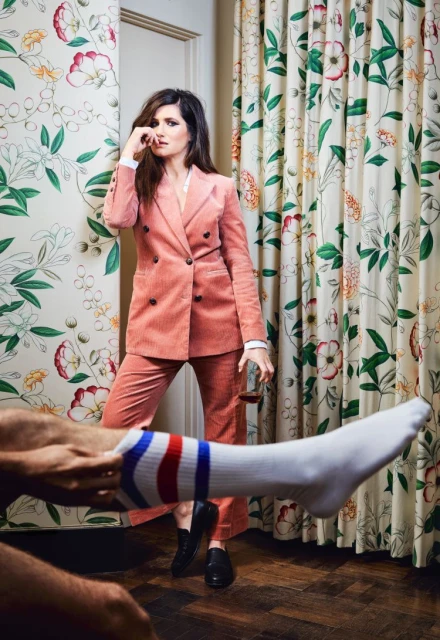 Woman in pink suit poses with glass as someone else puts on a sock in an indoor setting with floral wallpaper and curtains.
