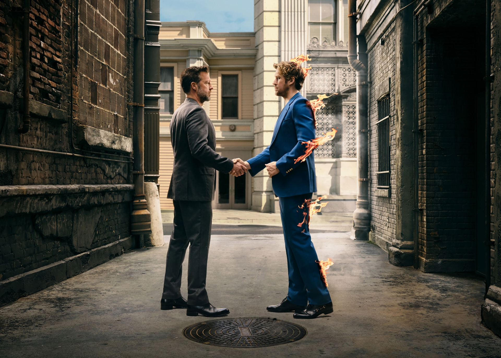 Two adult men in suits are shaking hands in a narrow alleyway, one engulfed in flames, in an eye-level medium shot.