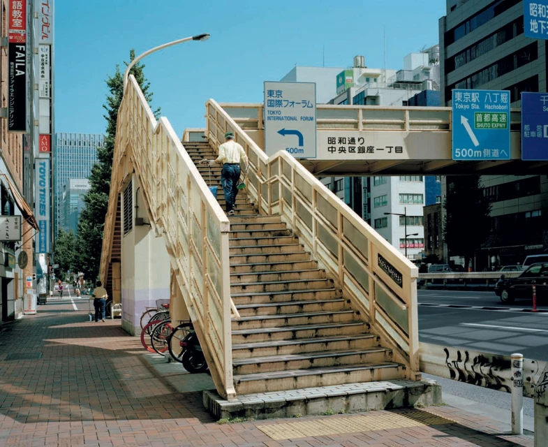 Urban scene captures a man ascending a pedestrian overpass in Tokyo, Japan, with city buildings and signs visible on a clear day.