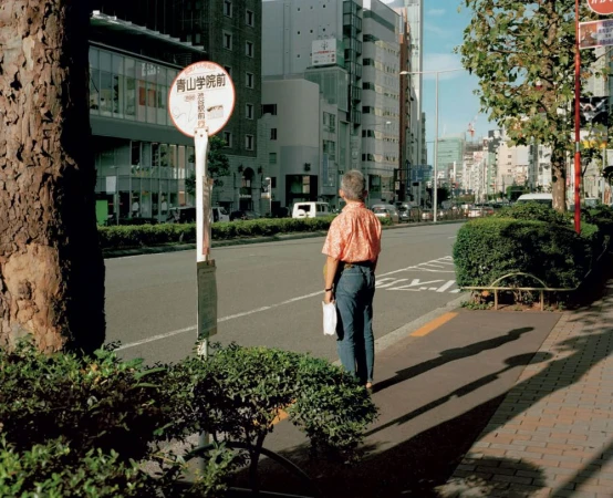 Mature adult stands at a bus stop in an urban street setting in daytime, wearing an orange shirt and blue jeans, looking toward the road.