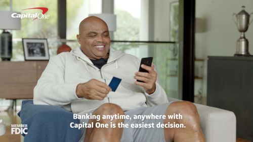 Charles Barkley sits in living room with a credit card and phone, smiling, in Capital One advertisement. Banking, finance, technology.