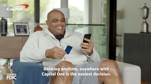 Charles Barkley sits in living room with a credit card and phone, smiling, in Capital One advertisement. Banking, finance, technology.