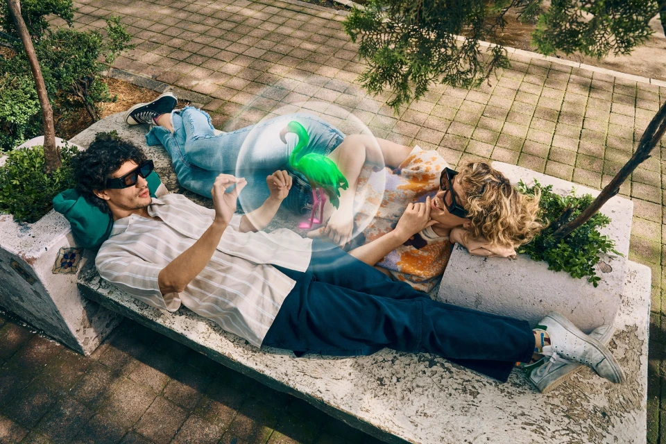 Two people wearing glasses are lying on a stone bench outdoors with plants nearby and a digitally created flamingo overhead.