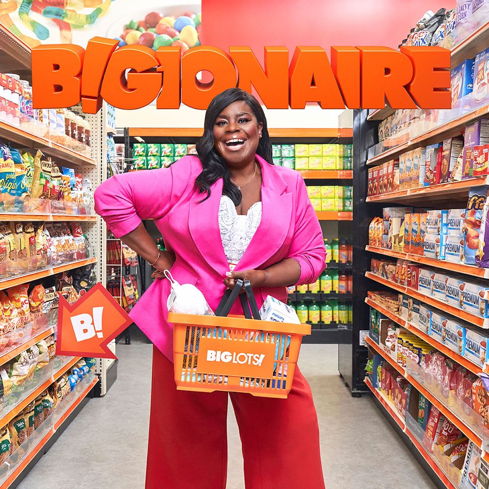 Smiling woman in a pink blazer holds an orange shopping basket in a grocery store aisle filled with snacks and pantry staples.