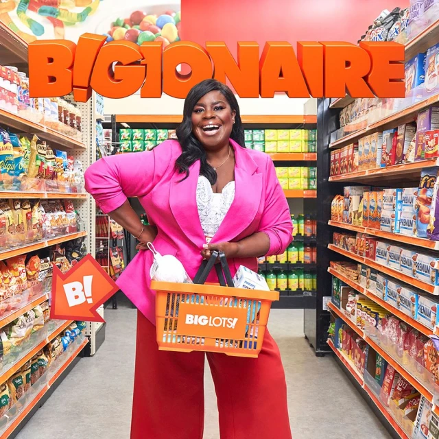 Smiling woman in a pink blazer holds an orange shopping basket in a grocery store aisle filled with snacks and pantry staples.