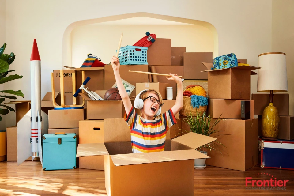 A child wearing headphones sits in a cardboard box playing drumsticks, surrounded by stacked boxes, a toy rocket, and house plants.
