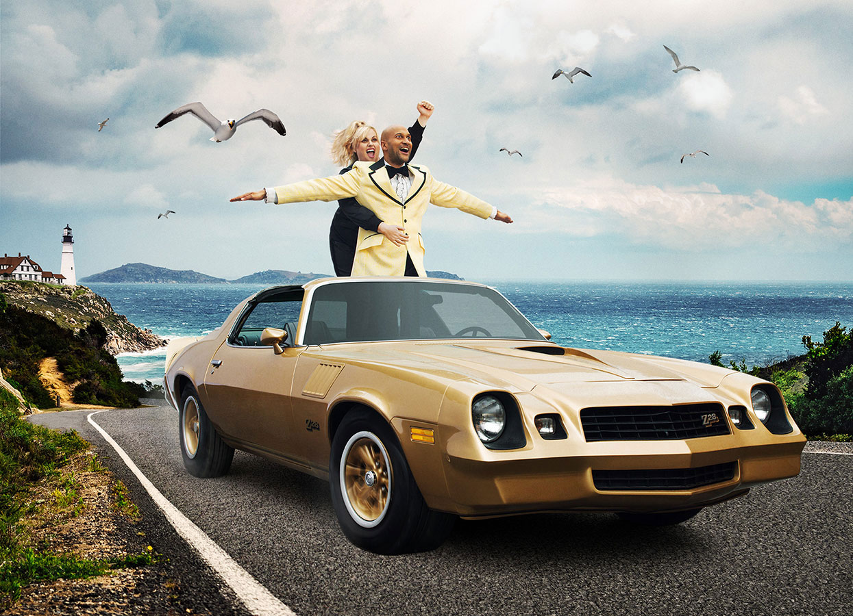 A couple stands in a gold convertible car with arms outstretched against a scenic coastal backdrop with blue ocean and scattered seagulls.
