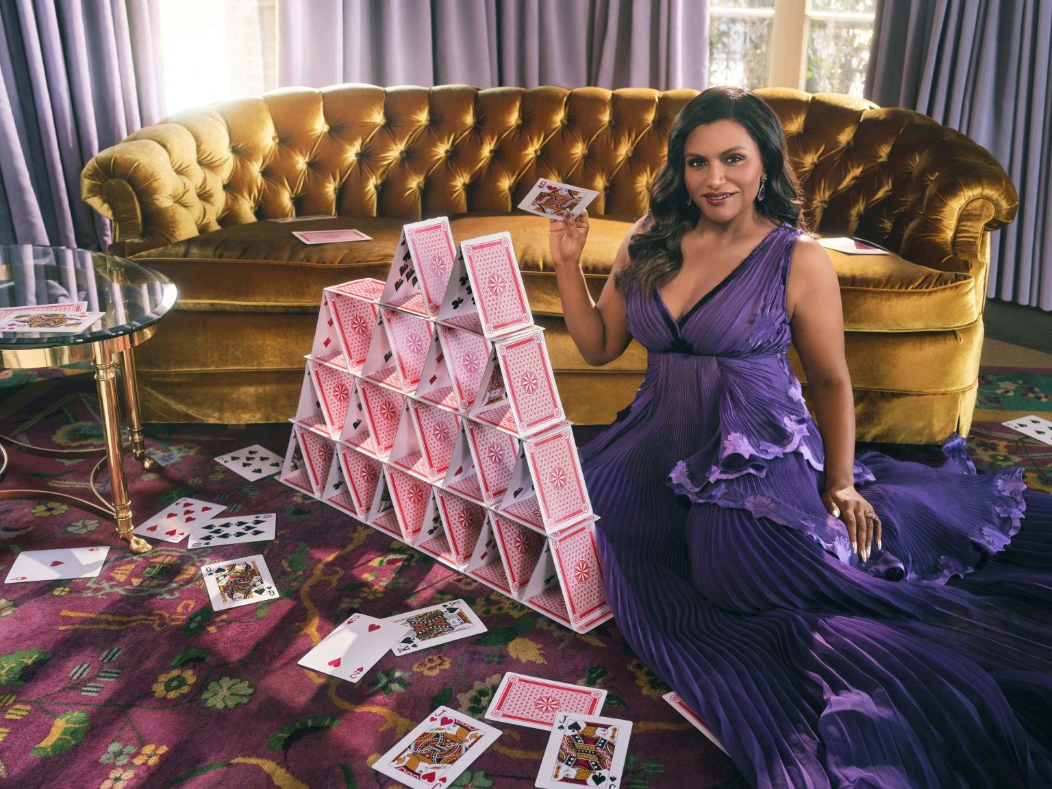 Adult woman in a purple dress sits on the floor next to a house made of playing cards, holding another card in her hand.
