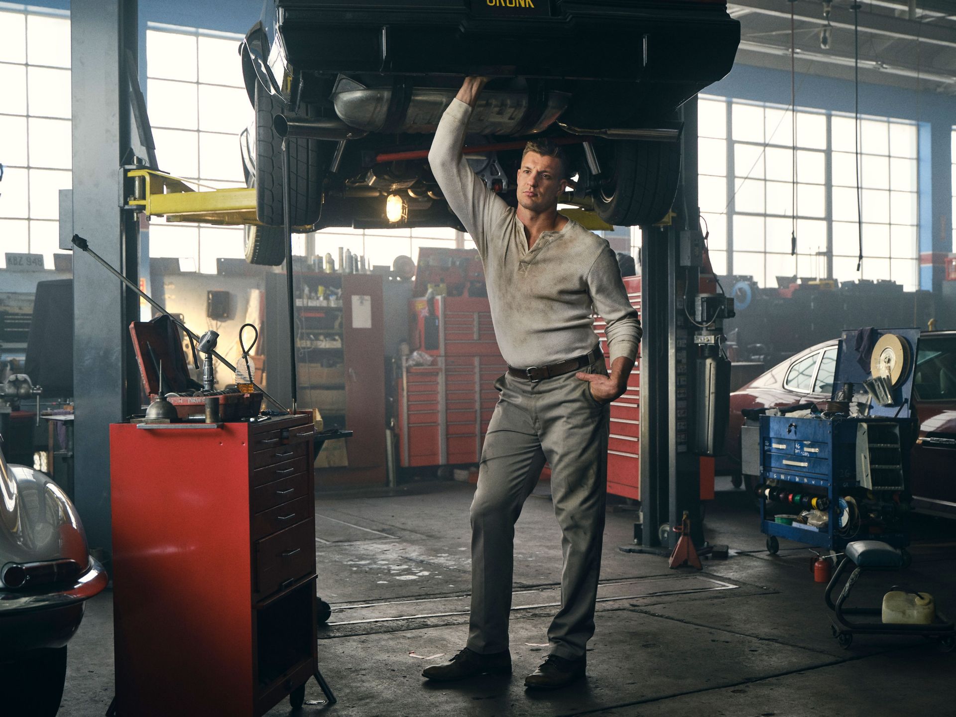 Mechanic working under a car in a garage, wearing a white shirt and grey pants, with a red tool chest nearby and cars in the background.