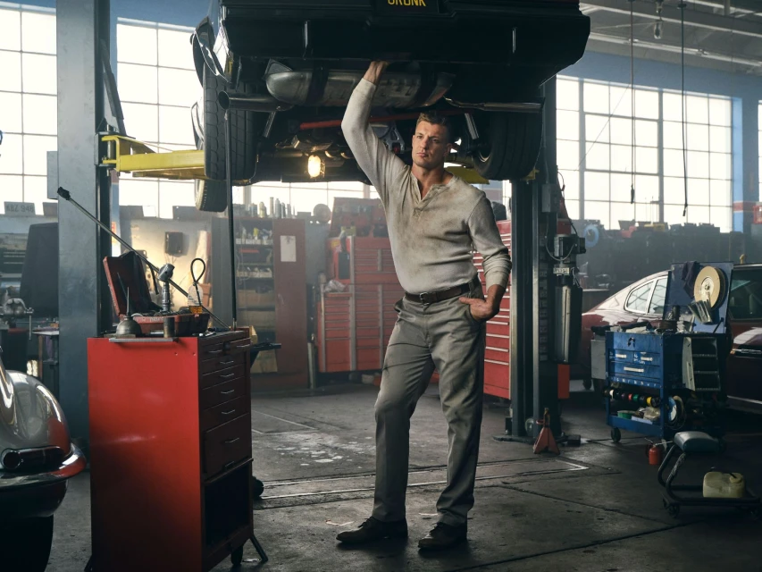 Mechanic working under a car in a garage, wearing a white shirt and grey pants, with a red tool chest nearby and cars in the background.
