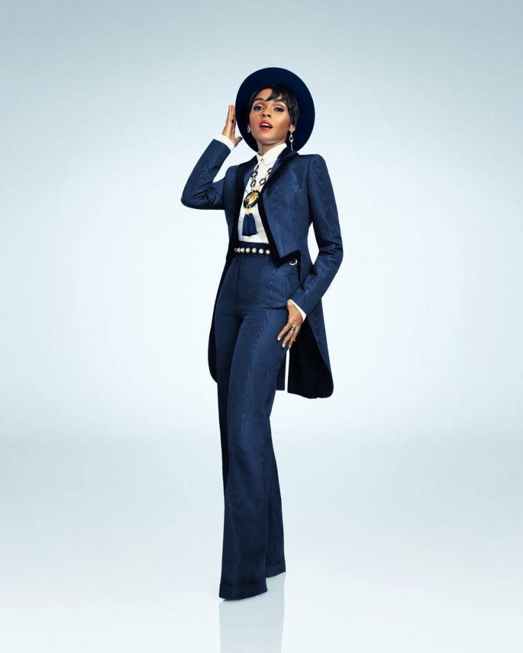 A striking portrait of an adult female fashion model in a blue suit and hat, standing confidently against a bright studio backdrop.