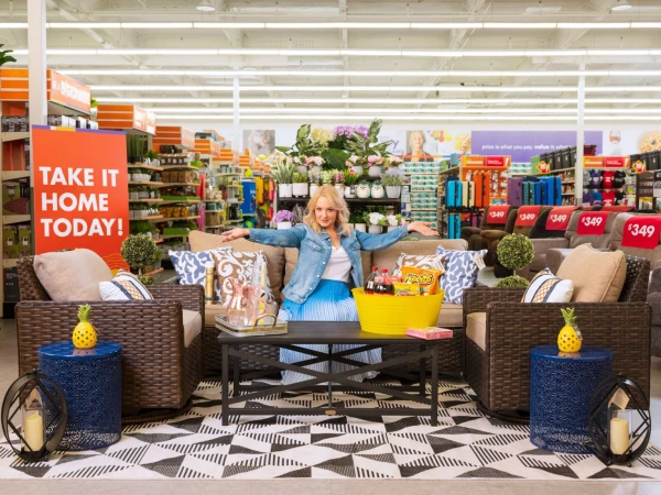 Woman poses on wicker patio furniture in a store display. Yellow tub with snacks rests on a coffee table, indoor store setting.
