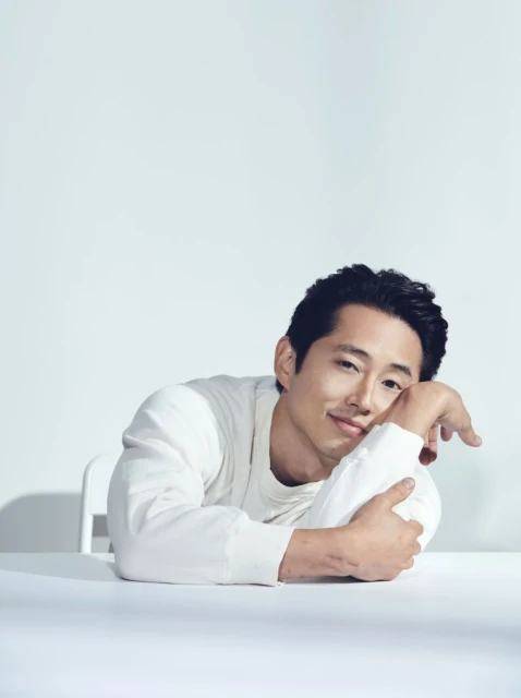 Headshot of a smiling Asian man with dark hair in a white sweater leaning on a white table against a light backdrop in a photo studio.