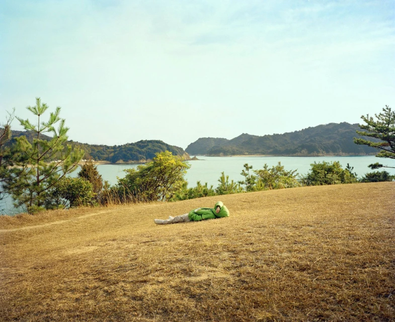 A lone person in a lime-green jacket lies on a grassy field overlooking a calm body of water with distant islands under a clear sky.