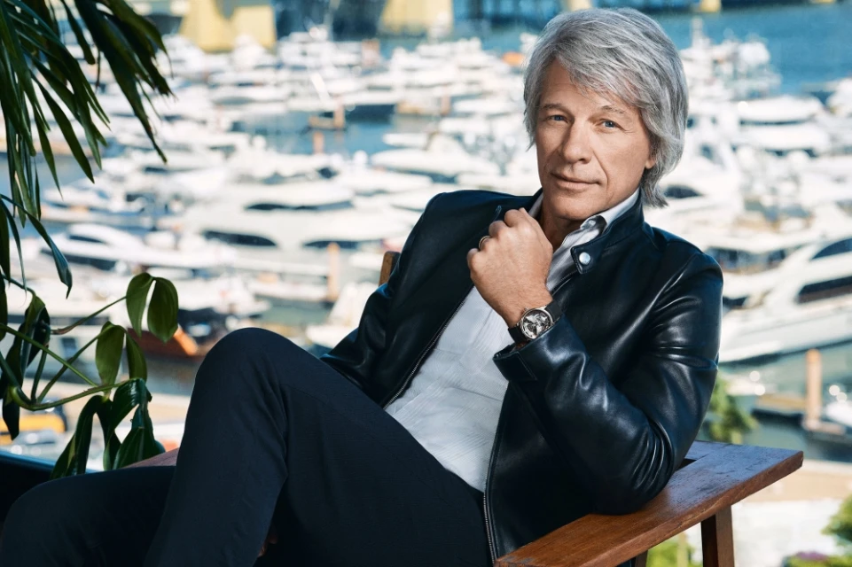 Portrait of adult male with grey hair wearing black leather jacket, white shirt, and watch, posing in front of harbor and yachts.