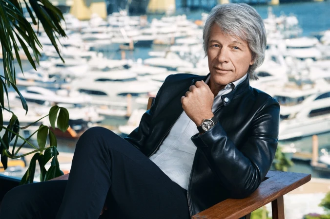 Portrait of adult male with grey hair wearing black leather jacket, white shirt, and watch, posing in front of harbor and yachts.
