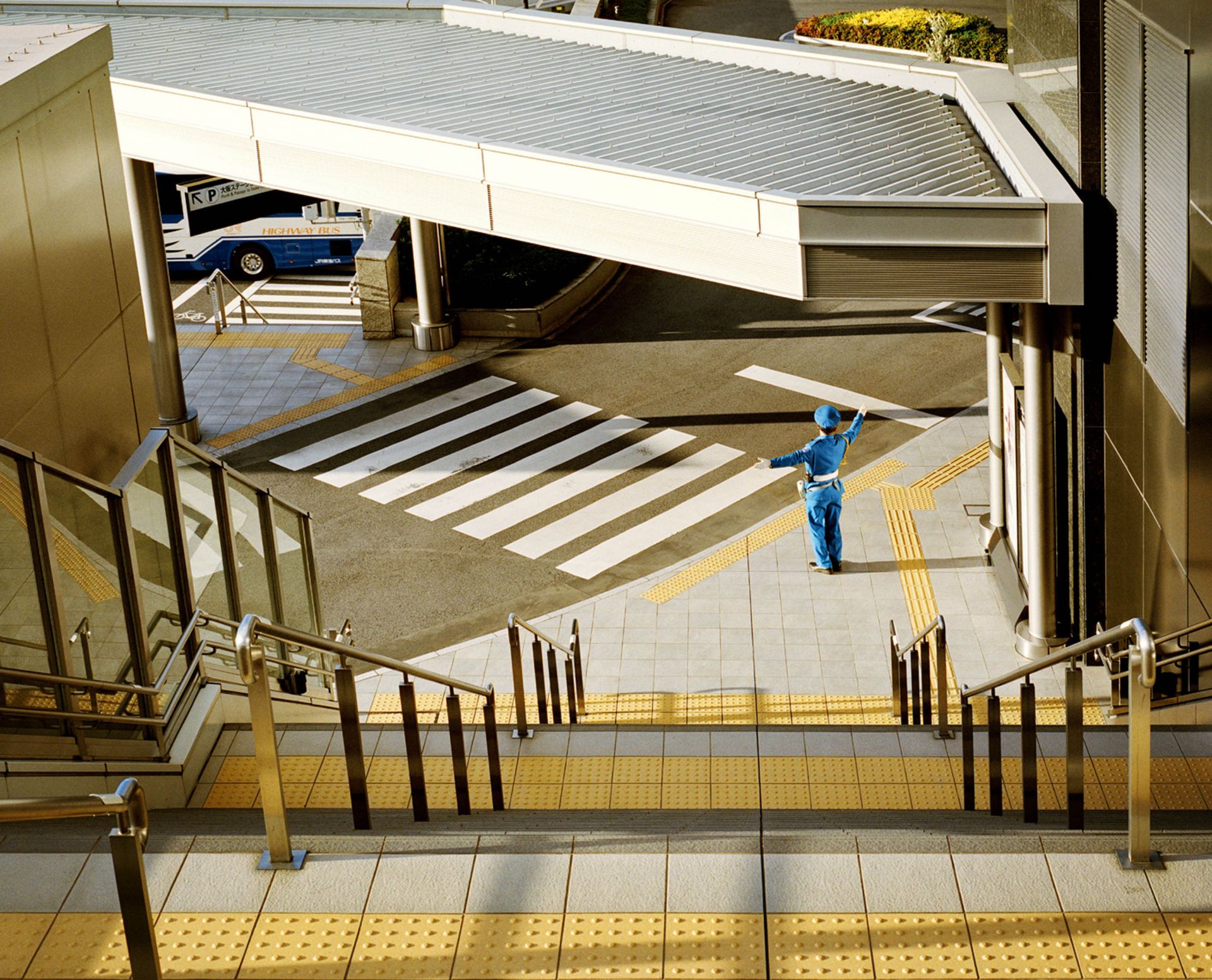 Traffic controller guides pedestrians at a zebra crossing on a city street, viewed from an elevated angle near a staircase with metal railings.
