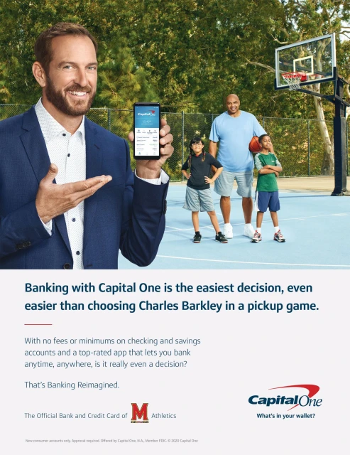 Smiling man in suit points to banking app on his phone with two kids and an adult near outdoor basketball court, Capital One ad.