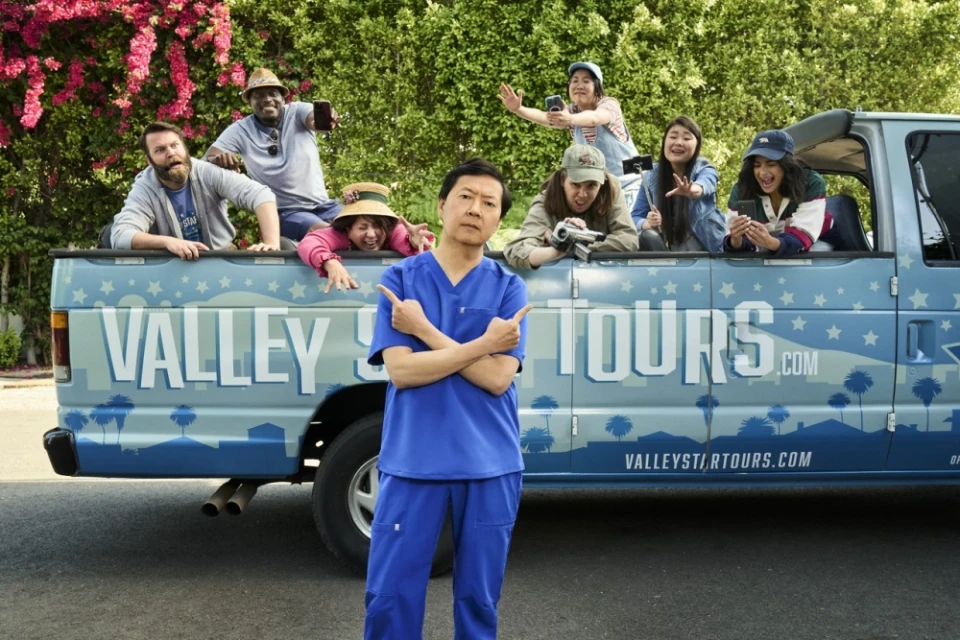 Promotional shot of Ken Jeong in scrubs in front of a van with tourists, taking photos against a leafy background in daylight.