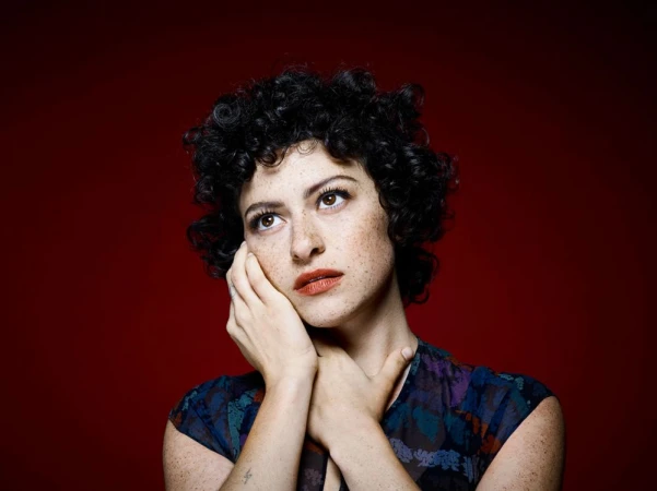 Portrait of a woman with dark curly hair and freckles posing against a dark red background by Emily Shur