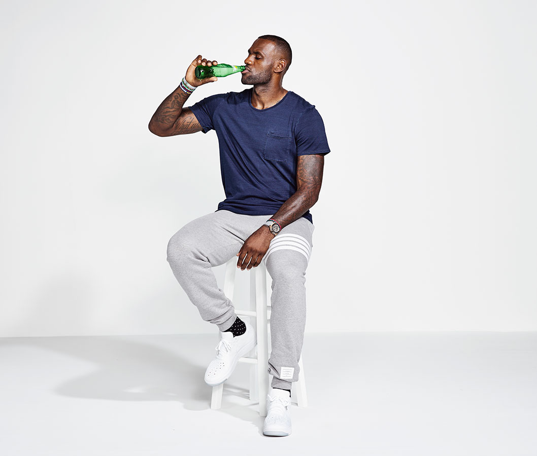 LeBron James drinks a green glass bottle while seated on a white stool in a light grey sweat suit and white sneakers, on white backdrop.