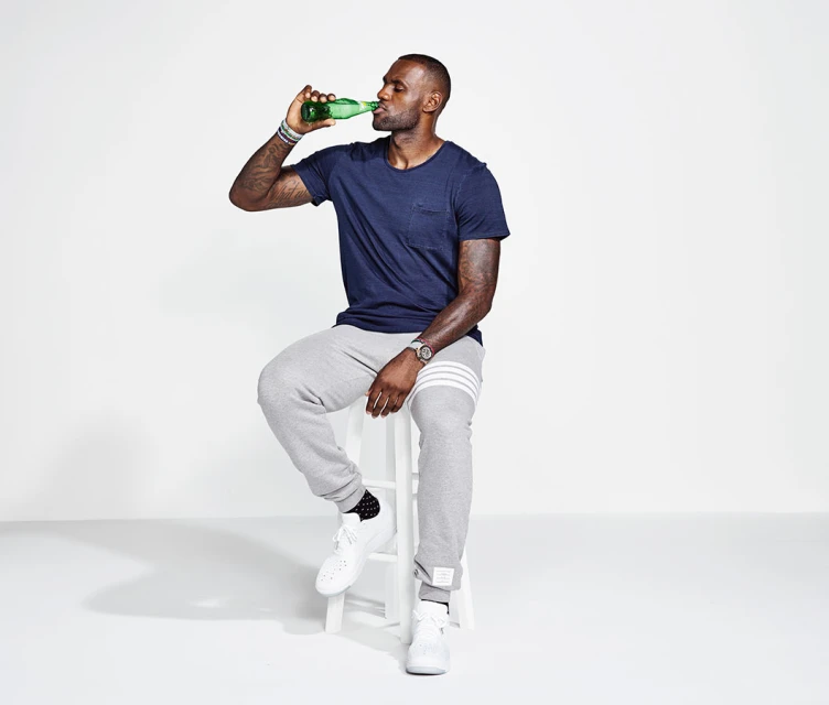 LeBron James drinks a green glass bottle while seated on a white stool in a light grey sweat suit and white sneakers, on white backdrop.