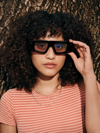 Portrait of a woman with dark curly hair wearing black sunglasses and a striped t-shirt against a textured tree bark background.
