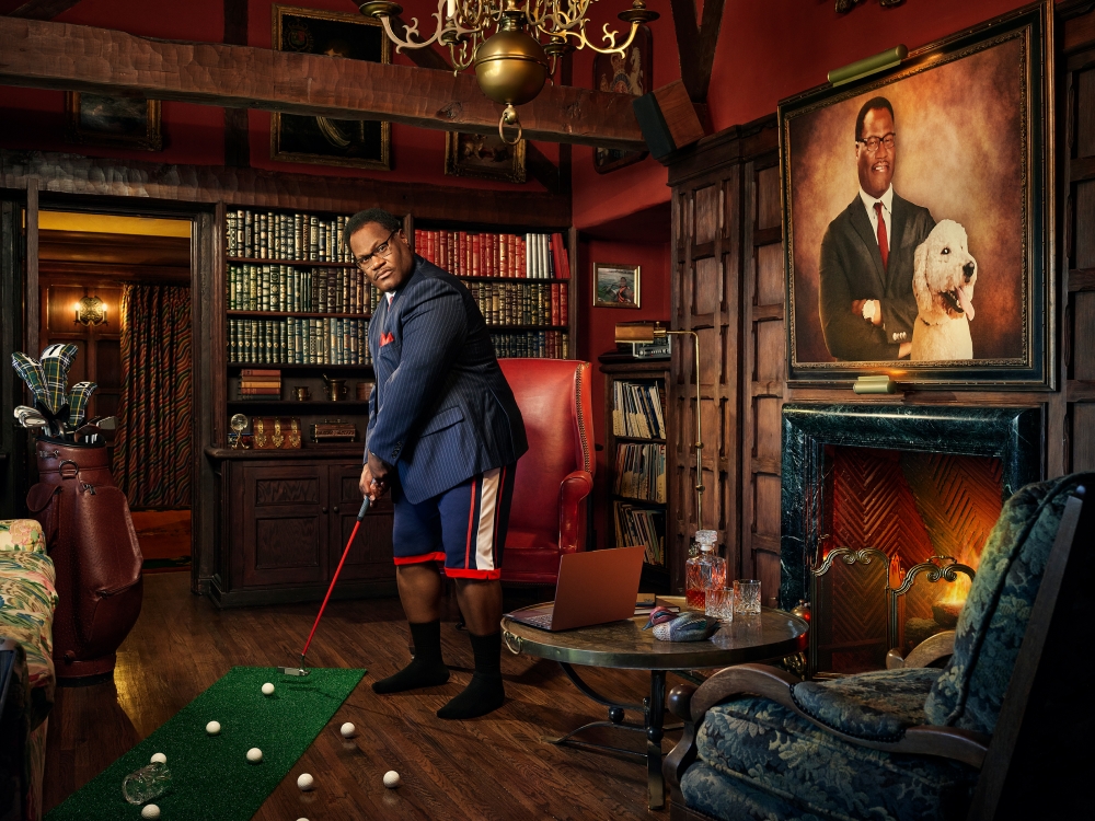 An adult male is indoors playing putt-putt golf in a darkly lit den, dressed in golf shorts and a blazer with books in the background.