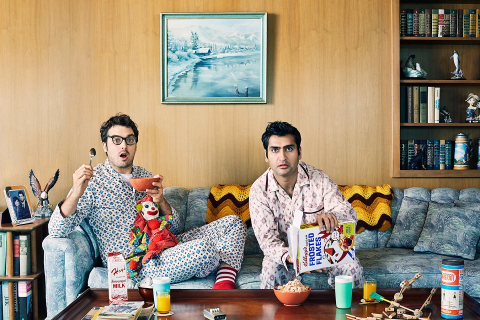 Two men in pajamas sit on a couch eating cereal, with a clown doll, Kellogg's Frosted Flakes, and vintage decor creating a retro living room scene.