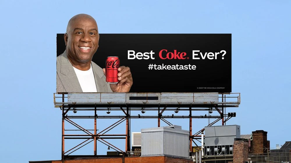 Billlboard features Magic Johnson holding a Coca-Cola, set against a clear blue sky with building tops below.