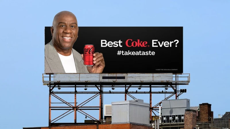 Billlboard features Magic Johnson holding a Coca-Cola, set against a clear blue sky with building tops below.
