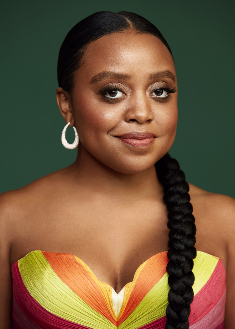 Portrait of a smiling African-American woman with braided hair, wearing a colorful strapless dress against a green backdrop.