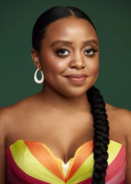 Portrait of a smiling African-American woman with braided hair, wearing a colorful strapless dress against a green backdrop.